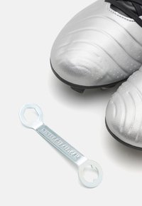 Botas de fútbol plateadas con superficie texturizada junto a una herramienta de llave de metal grabada con "nikefootball.com" sobre un fondo blanco.