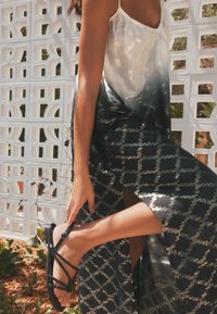 Sandales noires avec des lacets tressés complexes et un accent doré brillant, associées à une robe fluide et semi-transparente présentant un motif géométrique.