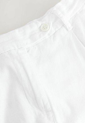 Witte linnen stof met een gladde textuur, voorzien van een sluiting met knopen aan de voorkant en netjes gestikte naden. De tailleband is gevouwen en licht gebogen.