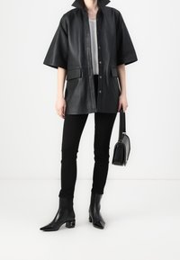 Chaqueta de cuero negra oversized con un alto cuello, dos bolsillos frontales, combinada con pantalones negros y botas de punta. Bolsa negra de mano.