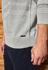 Pull gris clair avec des rayures texturées horizontales, des poignets et un ourlet côtelés. Il présente une petite étiquette d'accent noire en bas. Associé à un pantalon bleu marine.