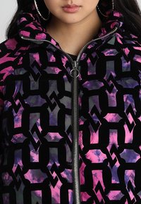 Pufferjack gemaakt van glanzende stof met een zwarte achtergrond en een levendig roze en paarse geometrische patroon, en een rits aan de voorkant.