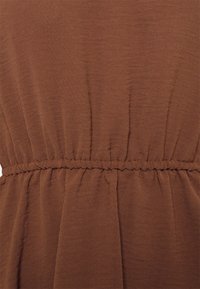 Tissu brun texturé avec une taille froncée. Tombé doux sous la taille, créant une silhouette fluide. Aucune motif ni élément métallique visible.