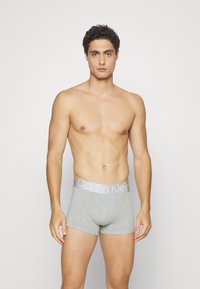 Sive pamučne bokserice s mekom teksturom, s kontrastnim svijetlo sivim pojasom s logotipom Calvin Klein. Model stoji, ruke uz tijelo.