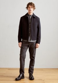 Joven hombre está de pie vistiendo chaqueta negra sobre camisa gris con botones, pantalones gris oscuro y zapatos de vestir negros sobre suelo de madera y fondo de pared lisa.
