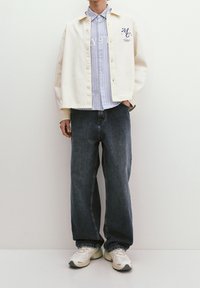 Cream button-up jacket over a light blue striped shirt, paired with dark baggy jeans and beige sneakers with black accents.