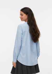 Chemise bleu clair à boutons, en tissu texteuré, avec un ourlet arrondi et des manches longues munies de poignets à boutons. Portée avec une jupe sombre.