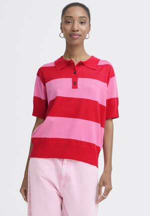 Woman wearing red and pink horizontal striped short-sleeve polo shirt and light pink pants, standing against a plain background.