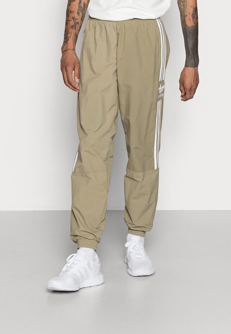 Pantalons coupe-vent beige avec des rayures blanches sur les côtés, taille élastique et chevilles resserrées, associés à des chaussures de sport blanches.