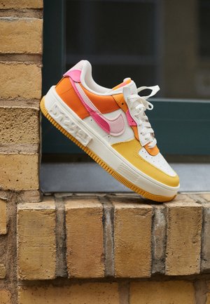 Basket basse avec des panneaux blancs, orange, jaunes et roses reposant sur un rebord en briques sur un fond sombre flou.