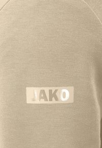 Felpa beige con una texture liscia e un logo rettangolare in argento lucido con la scritta "JAKO" sulla parte anteriore. Design semplice e minimalista.