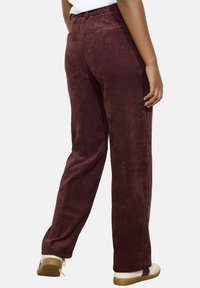 Persona che indossa pantaloni a gamba larga di velluto a coste bordeaux scuro e sneakers bianche, mostrata di spalle su uno sfondo semplice.