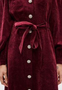 Burgundi színű corduroy ruha megkötős derékrésszel, díszített gombokkal, amelyeken virágos minta található, és hosszú ujjak. A textúrált szövet részletei jól láthatók.