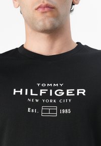 Zwarte katoenen t-shirt met ronde hals, voorzien van witte bedrukte tekst "TOMMY HILFIGER NEW YORK CITY Est. 1985" en een rechthoekig logoontwerp.