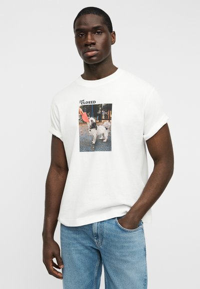 Λευκό βαμβακερό T-shirt με γυρισμένα μανίκια, με γραφικό εκτύπωσης ενός σκύλου και το κείμενο «Μείνε Κλειστός». Συνδυάζεται με μπλε τζιν παντελόνι.