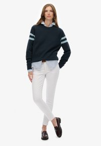 Marine gebreide trui met lichtblauwe strepen, gelaagd over een lichtblauw en wit gestreept shirt, gecombineerd met witte skinny broek en donkere instappers.