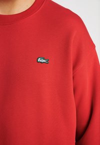 Felpa rossa con collo a girocollo, caratterizzata da un piccolo logo ricamato di un coccodrillo verde e bianco nella zona sinistra del petto.