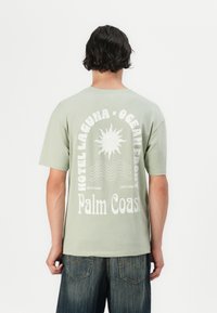 Lys grøn bomulds t-shirt med rund hals, der har hvid grafisk tekst og soldesign på ryggen, parret med mørke denim shorts.