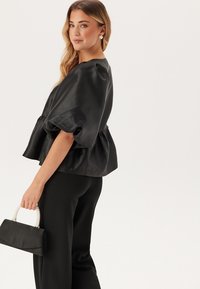Blusa di raso nera con maniche a sbuffo e vita arricciata, abbinata a pantaloni neri sartoriali e una piccola pochette nera con manico di perle.