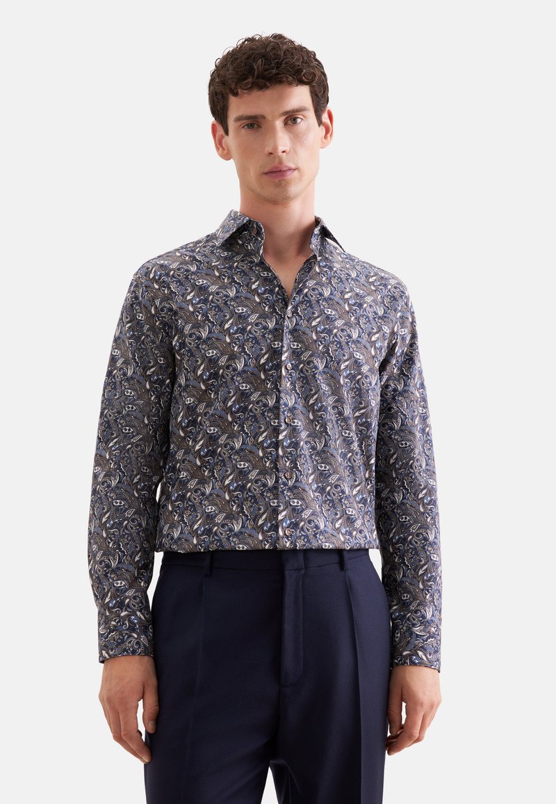 Homme portant une chemise à manches longues boutonnée avec un motif paisley bleu et blanc et un pantalon bleu foncé.