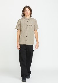 Kortærmet, beige-stribet skjorte med to brystlommer og sorte knapper, parret med sorte løstsiddende bukser og mørke sneakers.