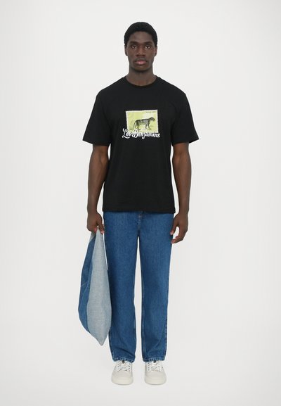 Μαύρο κοντομάνικο t-shirt με γραφικό print που απεικονίζει έναν δεινόσαυρο και κείμενο σε ανοιχτό φόντο, συνδυασμένο με μπλε τζιν και λευκά sneakers.