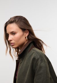 Giovane donna con lunghi capelli castani, che indossa una giacca verde scuro e orecchini a cerchio dorati, che guarda pensierosa a sinistra su uno sfondo neutro.