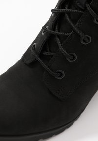 Svarta mockaboots med en slät yta, prydda med svarta hexagonala öglor och matchande snören. Designen är ankellång med minimal detaljering.