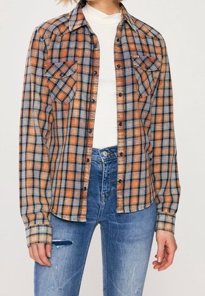 Mujer con camisa de cuadros naranja y azul desabrochada sobre un suéter blanco de cuello alto, combinada con jeans azules desgastados de tiro alto.