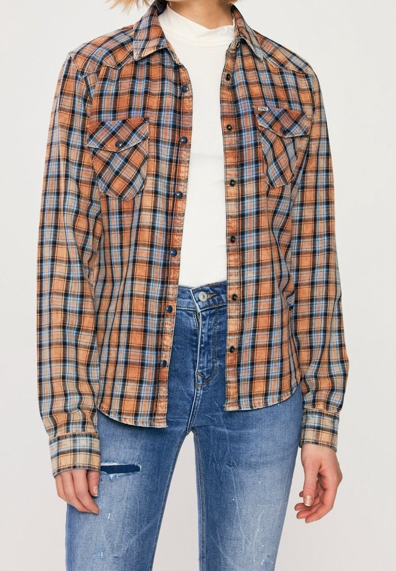 Femme portant une chemise à carreaux orange et bleue déboutonnée sur un col roulé blanc, associée à un jean bleu déchiré taille haute.