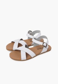 Sandales plates blanches avec des brides croisées et une boucle. Matériau doux et lisse, avec une semelle intérieure en cuir bronzé rembourrée et une semelle extérieure texturée.