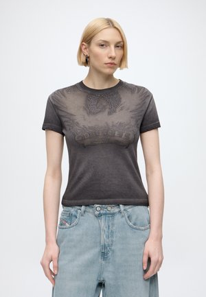 Giovane donna con capelli biondi corti che indossa una t-shirt nera effetto usato con stampa grafica e jeans larghi azzurri, in piedi davanti a uno sfondo semplice.