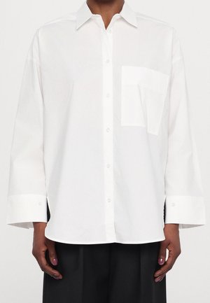 Osoba ubrana w luźną białą koszulę zapinaną na guziki z kieszenią z przodu, w połączeniu z czarnymi spodniami na jednolitym tle.