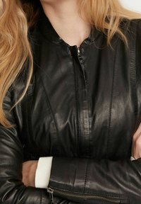 Svart skinnjacka med dragkedja framtill, ribbad textur och åtsittande silhuett. Har kontrastsömmar och vita ribbade mudd-detaljer.