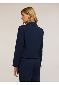 Blazer navy con vestibilità su misura, colletto a revers e maniche lunghe. La parte posteriore presenta un design pulito con dettagli minimali e cuciture strutturate.