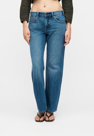 Nő, aki egyenes szárú kék farmert, rövid zöld kötött pulóvert és fekete, négyzet alakú orrú szandált visel, fehér háttér előtt áll.