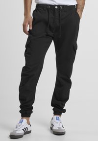 Man wearing black cargo joggers with elastic cuffs and white Adidas sneakers with black stripes and blue logos.
