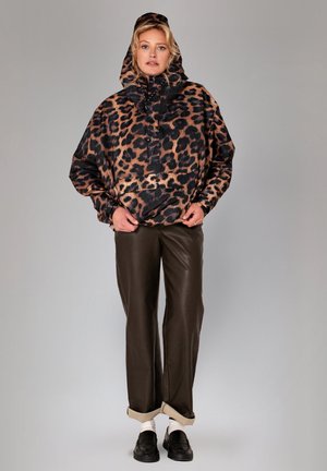 Vrouw draagt een capuchonjas met luipaardprint, donkerbruine broek met boorden, witte sokken en zwarte loafers, staand tegen een effen grijze achtergrond.