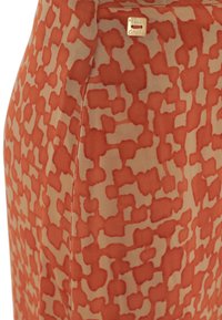 Tessuto a fantasia in arancione e beige, caratterizzato da un design astratto. Include un piccolo dettaglio con un logo dorato nella parte superiore.
