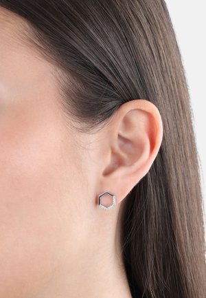 Pendiente de stud hexagonal plateado con superficie texturizada y pequeñas piedras preciosas transparentes a lo largo de un borde, llevado en la oreja izquierda.