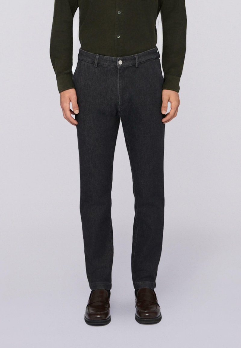 Uomo che indossa jeans slim-fit grigio scuro, camicia botton-down verde scuro e scarpe in pelle marrone scuro, in piedi contro uno sfondo chiaro.