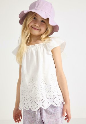 Ung pige smilende iført en hvid øjlet top med flæser på ærmerne, lyse lilla bukser med blomsterprint og en lyse lilla bøllehat.