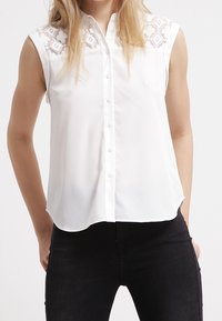 Blusa bianca senza maniche con abbottonatura frontale, caratterizzata da dettagli in pizzo sulle spalle e una texture di tessuto liscia e leggera.