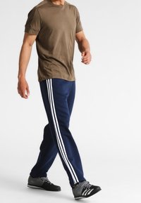 Olivgrünes Baumwoll-T-Shirt kombiniert mit marineblauen Jogginghosen, die drei weiße Streifen an den Seiten haben, getragen mit schwarzen und grauen Sportschuhen.
