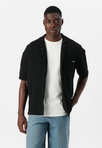 Schwarzes, strukturiertes Kurzarmhemd mit zwei Brusttaschen, über einem weißen T-Shirt getragen, kombiniert mit hellblauen Jeans. Minimale Markenkennzeichnung sichtbar.