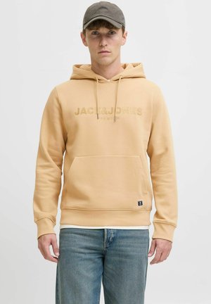 Beige huva sweatshirt med framficka, dragsko, broderad logotyp och ribbade muddärmar, parat med ljusblå jeans.