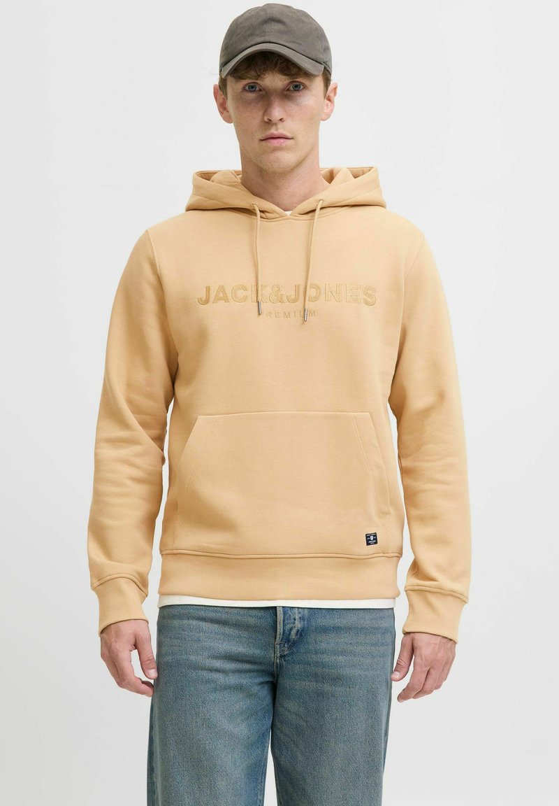 Sudadera con capucha beige, con bolsillo frontal tipo bolsa, cordones, logo bordado y puños de canalé, combinada con jeans azul claro.