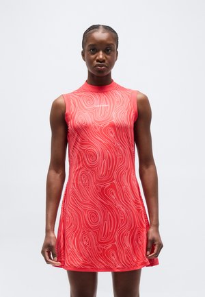 Žena v bezrukávových červených šatech s bílými abstraktními vzory ve tvaru spirál a logem "Calvin Klein", stojící před jednoduchým světlým pozadím.