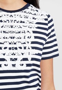 Laivan sininen ja valkoinen raidallinen paita, jossa on näyttävää valkoista pitsiä kaula-aukossa. Lyhyet hihat, pehmeä kangas, jossa on sileä rakenne.