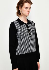 Schwarz-weißer Pullover mit strukturiertem, gemustertem Körper, schwarzem Kragen und drei goldenen Knöpfen an der Knopfleiste. Weiches Strickmaterial.